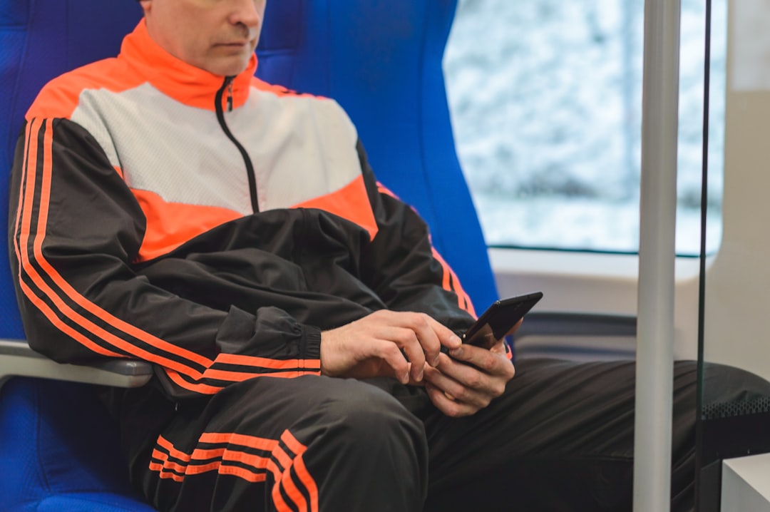 A man checking his smartphone by Virtual Personal Trainers man in orange and black jacket and black pants sitting on blue chair